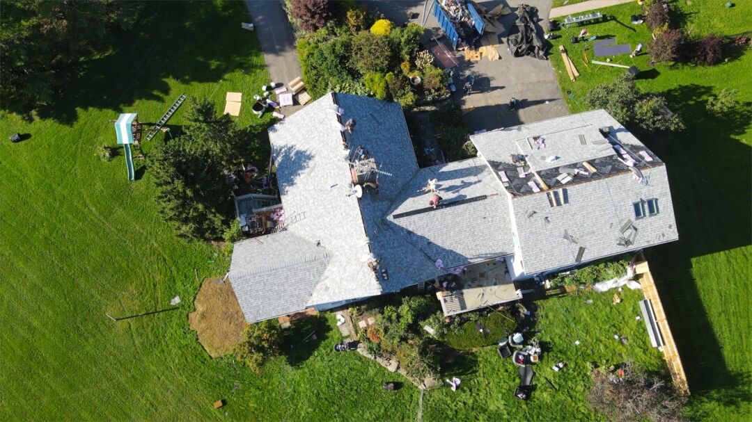 Aerial view of a large house with a complex, multi-level gray roof surrounded by green lawns, with construction materials and workers on the roof.