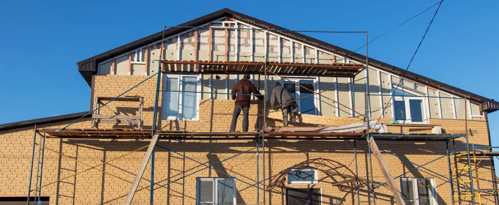 Two construction workers stand on scaffolding installing exterior siding on the upper level of a partially finished brick house under clear blue sky.