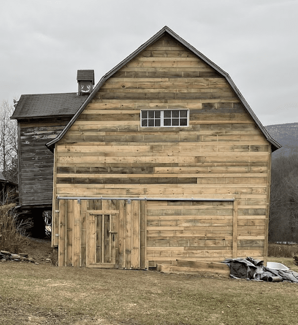 A wooden barn with a large sliding door and small window stands on grass; part of an older attached structure is visible to the left.