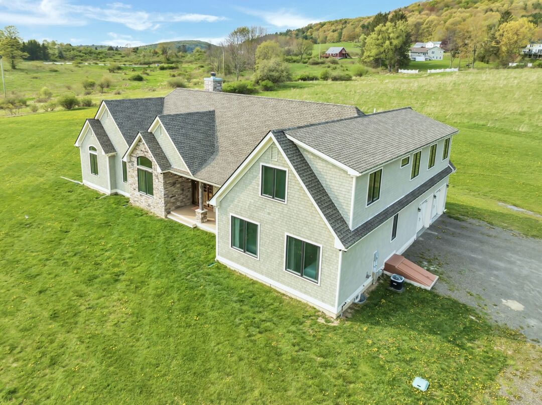 A large two-story house with light siding and multiple gables sits on a grassy property surrounded by rolling hills and scattered trees.