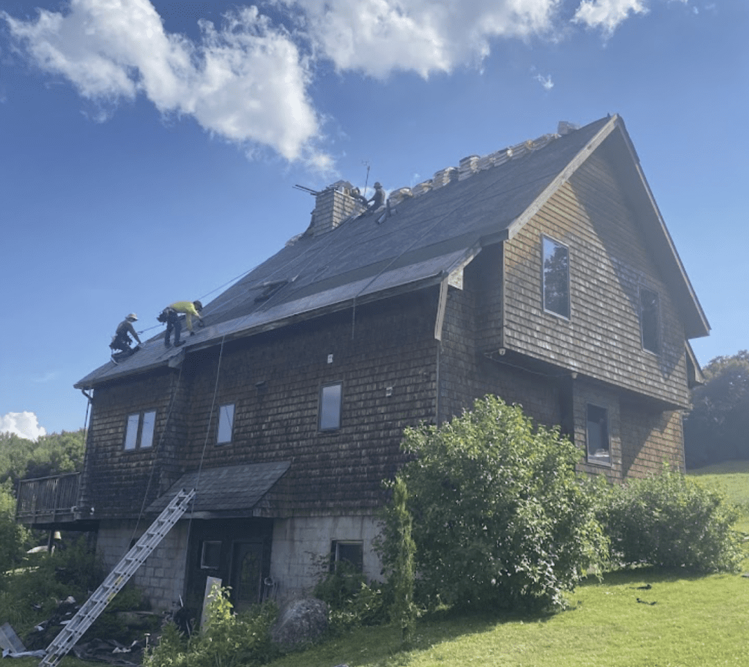 Three workers are repairing the roof of a wooden house on a sunny day, with ladders and construction materials visible on the roof.