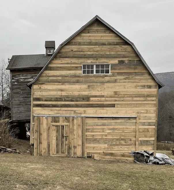 A wooden barn with a large sliding door and small window stands on grass; part of an older attached structure is visible to the left.
