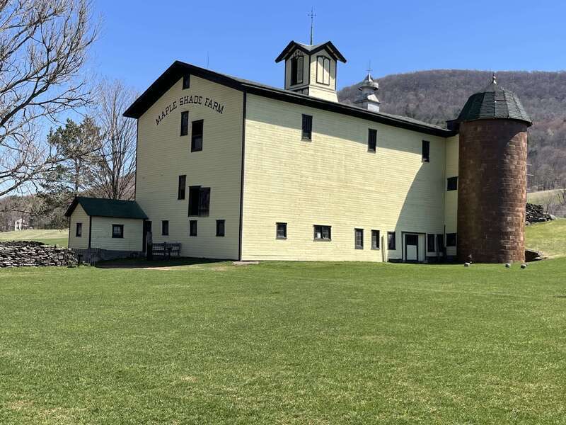 A large, pale yellow barn with a silo and cupola, labeled "Maple Shade Farm," stands on a grassy lawn with hills in the background.