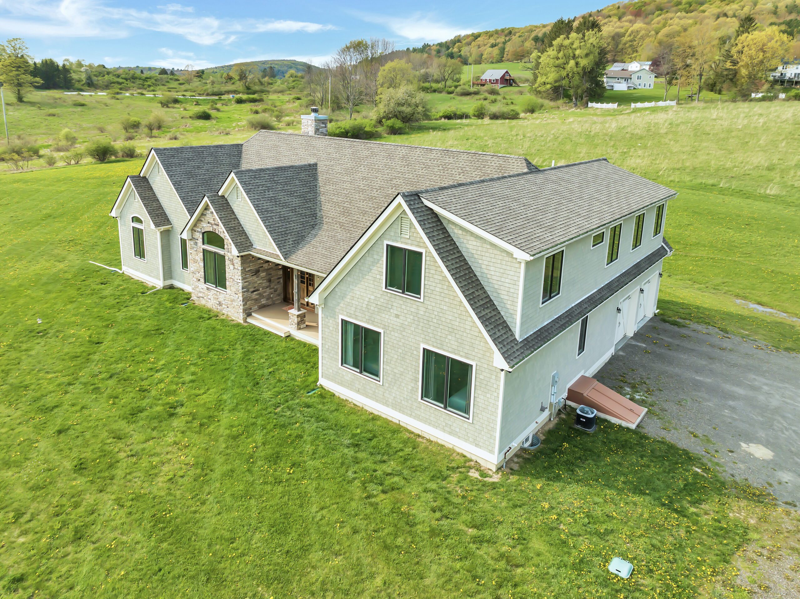 A large two-story house with light siding and multiple gables sits on a grassy property surrounded by rolling hills and scattered trees.