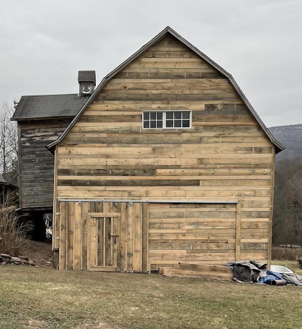 A wooden barn with a large sliding door and small window stands on grass; part of an older attached structure is visible to the left.