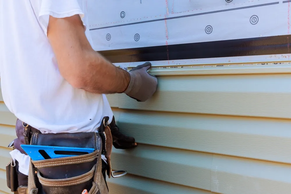 Person wearing gloves and tool belt installs light green horizontal vinyl siding on a house exterior wall.